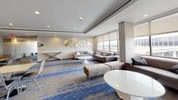 Located at 4400 East-West Hwy in Bethesda, Maryland, this polished single-floor commercial suite offers a thoughtfully designed layout suited for professional use. The open lounge area serves as the centerpiece of the space, furnished with contemporary gray sofas, accent ottomans, and sleek white coffee tables atop a bold blue and tan patterned carpet that runs throughout. Large windows with roller shades line the perimeter, flooding the space with natural light and offering panoramic views of the surrounding Bethesda cityscape.

A dedicated conference room provides a private setting for meetings, equipped with a wall-mounted large-format display, a soundbar, and a modest conference table with seating. The room features the same distinctive blue-toned carpet and clean white walls, creating a cohesive aesthetic.

The reception area features a smooth, white countertop desk framed by full-length mirrors that visually expand the space. Adjacent to it is a fully equipped kitchenette with light wood flooring, white cabinetry, stainless steel appliances including a full-size refrigerator and microwave, a wall oven, and a utility sink — ideal for supporting day-to-day office operations.

Throughout the suite, recessed lighting, subtle textured wall accents, and a neutral color palette contribute to a clean, professional ambiance.