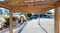 This covered carport and EV charging station is a single-level outdoor structure designed to serve the parking needs of a residential community. The structure features a sturdy wood pergola framework with open-slat beams that allow natural light to filter through, casting distinctive striped shadows across the paved surface below. Anchored by robust timber posts, the canopy provides shelter while maintaining an open, airy feel.

A dedicated electric vehicle charging unit is prominently positioned within the covered area, offering convenient on-site charging capability for residents. Adjacent to the charger, a freestanding waste receptacle adds practical utility to the space. The paved surface is marked with parking stall striping, clearly delineating individual spaces.

The surrounding landscape includes ornamental grasses and mature shrubs that border the structure, adding natural texture and visual interest. A wooded tree line frames the rear of the property, providing a green backdrop and a sense of natural seclusion. Views from within the carport reveal a multi-story residential building with balconies situated across the parking lot, indicating the broader community context. The overall setting is well-maintained, combining functional infrastructure with thoughtful landscaping in an open, accessible outdoor environment.