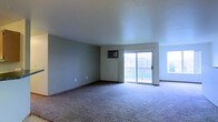 This single-floor space offers a straightforward, functional layout centered around a generously sized living area. The room is finished with wall-to-wall carpeting in a neutral gray-beige tone, providing a soft underfoot feel throughout. The walls are painted in a muted sage green, contributing to a calm and understated atmosphere.

A prominent sliding glass door serves as the focal point of the main living area, allowing a substantial amount of natural light to fill the room and providing direct access to an outdoor balcony. The balcony features a railing and offers views of the surrounding exterior environment, adding a sense of openness to the interior. An additional window positioned to the right of the sliding door further enhances natural light penetration.

A wall-mounted air conditioning unit is installed above the sliding door, offering climate control for the space. The ceilings are smooth and finished in white, maintaining a clean, uncluttered aesthetic. Electrical outlets are distributed along the baseboards, allowing for flexible furniture arrangement. Overall, the space presents a clean, move-in-ready environment with practical features suited for everyday living.