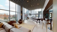 This upscale amenity floor is defined by its striking architectural design, combining soaring floor-to-ceiling glass curtain walls, a distinctive grid-pattern ceiling, and high-end finishes throughout. Abundant natural light pours in from multiple directions, offering panoramic city skyline views that serve as a dynamic backdrop across nearly every area of the floor.

The main lounge is an expansive, open gathering space anchored by plush seating arrangements, leather swivel chairs, and a shuffleboard table, all set atop artfully designed area rugs over large-format tile flooring. Sheer drapery softens the wall-to-wall glazing while maintaining the visual connection to the cityscape beyond. Sliding glass doors provide direct access to an outdoor terrace, extending the entertainment options outdoors.

A chef's kitchen features a dramatic stone slab accent wall, a substantial stone island with bar seating, integrated appliances, and rich wood cabinetry. Adjacent to this, a stylish bar area offers a marble-topped counter, open shelving, and barstool seating beneath warm pendant lighting.

The conference room accommodates a large group around a striking dark stone oval table, flanked by upholstered chairs and floor-to-ceiling windows. Glass-enclosed private offices and collaborative work areas line a wood-paneled corridor. The entry lobby presents dark marble columns, a curved reception desk, and refined lounge seating, setting an elegant tone upon arrival.