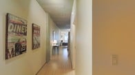 This thoughtfully designed single-floor space blends industrial and contemporary aesthetics to create a distinctive and inviting environment. The hallway entry sets the tone immediately, showcasing an exposed concrete ceiling that contrasts beautifully with the warm-toned hardwood flooring running throughout. Clean white walls provide a neutral backdrop that highlights curated decorative touches, including eclectic wall art that adds personality and character to the space.

The corridor leads naturally toward a bright, open living area visible at the end of the hall, where natural light pours in through generously sized windows, creating an airy and welcoming ambiance. A slim console table with a lamp provides a tasteful accent along the hallway, offering both function and style.

The living area beyond features seating and furnishings that complement the modern industrial feel of the space, with the natural light further enhancing the sense of openness. The seamless flow from the entry corridor into the main living area gives the space a cohesive and well-considered layout. Overall, the combination of raw concrete, warm wood tones, abundant natural light, and carefully chosen décor creates a space that is both visually striking and comfortably livable.