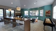 This single-story space offers a thoughtfully designed interior that blends functionality with contemporary style. The room is defined by striking teal accent walls that contrast with neutral gray tones, creating a visually dynamic environment. Elevated ceilings contribute to an open, airy atmosphere, while recessed lighting and a sculptural geometric pendant light fixture serve as a focal point, adding artistic flair to the space.

A large wooden communal table with modern bar-height seating anchors one side of the room, providing a practical workspace or social gathering area. Adjacent to it, a workstation area equipped with computer monitors offers a dedicated zone for productivity. On the opposite side, a comfortable lounge area features contemporary armchairs and sofas arranged around a central space, ideal for relaxed conversation or informal meetings.

Multiple windows along the teal-accented wall allow natural light to fill the room while offering views of the surrounding outdoor greenery. Patterned carpeting adds texture underfoot and ties the design elements together. The overall ambiance is modern, welcoming, and purposefully arranged to support both collaborative and individual use.