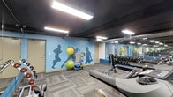 This single-floor fitness space is designed to support a well-rounded workout experience. The room is immediately distinguished by its bold, light blue accent wall adorned with dynamic silhouette murals of athletes in motion, creating an energizing atmosphere that encourages activity. A large wall-to-wall mirror runs along one side of the space, visually expanding the room and allowing users to monitor their form during exercise.

The flooring consists of durable, patterned carpet tiles in neutral grey tones, providing a practical and comfortable surface underfoot. Overhead, the dark-painted ceiling features exposed ductwork and flush rectangular panel lights, giving the space an industrial yet functional aesthetic.

Equipment is thoughtfully arranged throughout the room. Cardio machines, including what appear to be rowing or recumbent units, are positioned toward the front, while a well-stocked dumbbell rack lines the mirrored wall. A fitness equipment cart holds stability balls, resistance tools, and a yoga mat, offering versatility for a variety of training styles. Posted fitness center rules near the doorway reflect an organized and user-friendly environment.