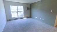 This single-floor space presents a straightforward and functional interior. The room visible is finished with soft, light gray wall-to-wall carpeting that lends a neutral, comfortable feel underfoot. The walls are painted in a muted sage green tone, creating a calm and understated ambiance throughout the space.

A large double-hung window dominates one wall, allowing an abundance of natural light to fill the room. The window offers a view of an outdoor green area and neighboring structures, providing a sense of connectivity to the surrounding environment. A baseboard heating unit runs along the wall beneath the window, ensuring efficient climate control during cooler months.

Complementing the heating system, a wall-mounted air conditioning unit is installed on the adjacent wall, offering year-round temperature regulation. Multiple electrical outlets are positioned along the lower portion of the walls, providing practical accessibility throughout the room.

The overall design is clean and minimalist, with smooth walls and no ornate detailing, making the space versatile and well-suited for a variety of uses. The combination of ample natural light, neutral tones, and functional climate systems contributes to a livable and practical environment.