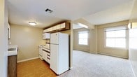 This single-floor space features an open-concept layout that seamlessly connects the kitchen and living area. The kitchen is equipped with white appliances, including a full-size refrigerator, electric range, and microwave, complemented by wood-trimmed upper cabinetry and white lower cabinets offering ample storage. Hardwood flooring grounds the kitchen area, while the adjacent living space transitions to wall-to-wall carpeting, creating a clear yet flowing distinction between the two zones. A structural column subtly defines the boundary between the kitchen and living area without disrupting the open feel. The living area benefits from natural light streaming through two windows fitted with white roller shades, contributing to a bright and airy atmosphere. Neutral beige walls and white ceilings run throughout the space, providing a clean, understated backdrop. Overhead ventilation is provided by a ceiling-mounted HVAC vent, and recessed and surface-mounted lighting ensures consistent illumination. The overall layout is functional and straightforward, making efficient use of the available floor space.