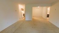 This single-floor space offers a straightforward, functional layout designed for comfortable everyday living. The main living area is finished with wall-to-wall neutral-toned carpeting that extends throughout, creating a cohesive and warm feel underfoot. White walls provide a clean, bright backdrop, while flush-mount ceiling lighting ensures consistent illumination across the space.

A partial dividing wall separates the main living area from an adjacent open room, which shares the same carpeted flooring and white walls. This secondary space features a visible ceiling vent, suggesting a dedicated climate control system, and benefits from the same overhead lighting style.

Through a doorway visible from the main living area, the kitchen is accessible and clearly defined as a separate room. It features wood-toned upper cabinetry, a white refrigerator, and a dark-finished range, providing essential cooking and storage functionality. The kitchen appears to have hard flooring, contrasting with the carpeted living spaces.

Overall, the space presents a neutral, move-in-ready environment with a simple, open feel that allows for flexible furniture arrangement and personalization.