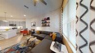 Located in Tampa's dynamic Channel District at 1251 Ray Charles Blvd, this single-floor residence showcases a thoughtfully curated interior that balances contemporary design with warm, eclectic character. The open-concept living area is anchored by a white-painted exposed brick accent wall adorned with floating wood shelves displaying vinyl records, lending a creative, music-inspired atmosphere. A generous sectional sofa, colorful area rug, and natural wood coffee tables create a relaxed yet stylish setting, while a ceiling fan and large windows with patterned drapes allow ample natural light and access to an outdoor area. The kitchen is well-appointed with sleek gray cabinetry, stainless steel appliances including a full-size refrigerator, electric range, and microwave, all complemented by a crisp white quartz countertop and a peninsula with bar seating beneath pendant lighting. Light wood-tone flooring runs throughout the main living areas, adding warmth and continuity. The bedroom features deep navy blue accent walls, plush carpeting, large windows, a dedicated workspace, and decorative shelving. The bathroom continues the navy and gray palette with wood-look flooring, a white vanity with quartz countertop, and a glass-enclosed shower, completing this well-designed Tampa residence.