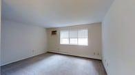 This single-floor, one-bedroom, one-bathroom unit offers a functional and comfortable living environment. The main living area is generously proportioned, featuring wall-to-wall carpeting that adds warmth underfoot. Neutral-toned walls and a clean white ceiling create a bright, airy atmosphere throughout the space. A large multi-pane window dressed with horizontal blinds allows natural light to flow into the room while providing privacy control. Climate comfort is well-addressed with both a wall-mounted air conditioning unit and a baseboard heating system, ensuring a pleasant environment across all seasons. The straightforward layout makes efficient use of the available space, offering a practical and livable floor plan. Multiple electrical outlets are positioned along the walls, providing convenient access for furnishings and electronics. The overall design is clean and neutral, offering a blank canvas that can easily be personalized to suit a variety of tastes and lifestyles. This unit is well-suited for individuals or couples seeking a comfortable, low-maintenance living space with essential amenities already in place.