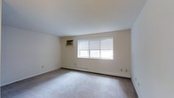 This single-floor, one-bedroom, one-bathroom unit offers a functional and comfortable living environment. The main living area is generously proportioned, featuring wall-to-wall carpeting that adds warmth underfoot. Neutral-toned walls and a clean white ceiling create a bright, airy atmosphere throughout the space. A large multi-pane window dressed with horizontal blinds allows natural light to flow into the room while providing privacy control. Climate comfort is well-addressed with both a wall-mounted air conditioning unit and a baseboard heating system, ensuring a pleasant environment across all seasons. The straightforward layout makes efficient use of the available space, offering a practical and livable floor plan. Multiple electrical outlets are positioned along the walls, providing convenient access for furnishings and electronics. The overall design is clean and neutral, offering a blank canvas that can easily be personalized to suit a variety of tastes and lifestyles. This unit is well-suited for individuals or couples seeking a comfortable, low-maintenance living space with essential amenities already in place.