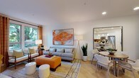 This single-floor residence offers an open-concept layout that seamlessly connects the living room, dining area, and kitchen into one cohesive, light-filled space. The living room serves as the focal point, anchored by a contemporary gray sofa paired with a vibrant orange upholstered ottoman and complementary accent stools. A large abstract painting in bold orange and white tones adds a striking artistic element to the neutral white walls. Recessed lighting and a tall floor lamp provide layered illumination, while a large window fitted with patterned drapes invites generous natural light and views of lush greenery outside. Light-toned hardwood flooring runs throughout, enhancing the airy, warm atmosphere.

Adjacent to the living area, the dining space accommodates a round table with wicker-style chairs, offering an intimate setting for meals. A large mirror on the wall amplifies the sense of space. Beyond, the kitchen is visible and features pendant lighting, modern appliances, and a clean, bright design. The overall ambiance is contemporary, welcoming, and thoughtfully styled with a cohesive warm color palette.
