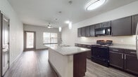 This well-appointed single-floor residence offers a thoughtfully designed open-concept layout that seamlessly connects the kitchen, dining, and living areas. The kitchen is a standout feature, showcasing rich dark espresso cabinetry with sleek chrome hardware, granite countertops, and a large central island with a built-in sink — all illuminated by elegant pendant lighting. Black appliances, including a range and microwave, complement the stainless steel refrigerator, while under-cabinet lighting adds a warm, functional glow. Wood-look flooring runs throughout the main living area, enhancing the modern aesthetic.

The living space opens toward a glass door and windows that invite natural light and offer views of the surrounding exterior. A ceiling fan provides added comfort to the airy, open layout. The bedroom features plush carpeting, neutral tones, and a well-sized window with exterior views, creating a calm and restful atmosphere. The bathroom is clean and functional, featuring a soaking tub with tiled surround, wood-look flooring, dark cabinetry for storage, and granite countertop accents. Together, these spaces deliver a cohesive, contemporary living experience with quality finishes at every turn.