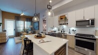 Located at 2600 N Orange Ave in Orlando, Florida, this single-floor residence offers a thoughtfully designed open-concept layout with contemporary finishes throughout. The kitchen serves as the heart of the home, boasting a large center island with seating, crisp white quartz countertops, a classic subway tile backsplash, and a mix of white and natural wood cabinetry. Stainless steel appliances, a farmhouse-style sink with matte black fixtures, and glass globe pendant lights complete the well-equipped space, which flows seamlessly into the living area. The primary bedroom features tray ceilings, wood-look flooring, large windows with draping curtains, and a ceiling fan, creating a calm and restful retreat. A second bedroom offers an earthy, cozy aesthetic with sage green accent walls and natural wood furnishings. Both bedrooms are served by bathrooms finished with light wood vanities, quartz counters, and wood-look tile floors—each leading directly into a dedicated walk-in closet with open shelving systems. One bathroom features a soaking tub, while the other offers a streamlined layout with bold deep green accent walls. Wood-look flooring ties the entire residence together, contributing to a warm, modern ambiance throughout.