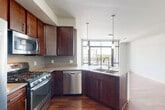 This single-floor, one-bedroom, one-bathroom residence offers a thoughtfully designed layout that maximizes functionality and style. The open-concept kitchen and living area serves as the heart of the home, anchored by rich dark-wood cabinetry, granite countertops, and a full suite of stainless steel appliances including a gas range, built-in microwave, dishwasher, and refrigerator. A peninsula with a double undermount sink provides additional prep space and casual seating, while pendant lighting adds a polished finishing touch. Large floor-to-ceiling windows flood the living area with natural light and frame expansive urban views, with sliding glass doors opening directly onto a private balcony — an ideal spot for taking in the surrounding cityscape. Rich hardwood flooring runs throughout the main living spaces, lending warmth and continuity. The bedroom features neutral-toned carpet and is a comfortable, quiet retreat. The bathroom is finished with a dark wood vanity, white countertop, undermount sink, and a framed mirror, all set against light tile flooring and neutral walls. A hallway connects the spaces efficiently, completing this well-organized urban residence.