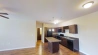 This two-story residence offers a thoughtfully designed layout spread across two levels. The kitchen serves as a central highlight, showcasing rich dark wood cabinetry that extends to the ceiling, complete with a built-in wine rack for added character. A center island with granite countertops provides generous prep space and a natural gathering point, while pendant lighting hangs above, adding warmth and visual interest. The kitchen is fully equipped with black appliances, including a refrigerator, range, microwave, and dishwasher, alongside a double sink set beneath additional counter space. Warm hardwood flooring flows seamlessly throughout the open-concept main living area, enhancing the inviting ambiance. The space is bathed in soft, even lighting from recessed fixtures and decorative pendants, creating a bright and welcoming atmosphere. The open sightlines between the kitchen and adjacent living areas foster a sense of spaciousness and connectivity. A hallway visible from the kitchen leads further into the home, hinting at the additional rooms and amenities that the two-floor layout provides. Clean wall finishes and generous ceiling height contribute to the overall airy, well-appointed feel of this residence.
