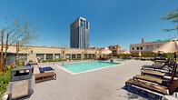 This thoughtfully designed outdoor amenity space offers a compelling blend of urban energy and relaxed leisure. At its heart is a generously sized rectangular swimming pool with clear turquoise water, framed by a wide, open sun deck furnished with lounge chairs and wooden side tables — ideal for sunbathing beneath open skies. The pool area is surrounded by low-rise brick and stone-clad building wings that lend the space a grounded, architectural character, while a dramatic high-rise tower visible on the horizon reinforces the downtown urban setting.

Encircling the central pool zone are elevated terrace walkways lined with black steel railings and raised planter beds brimming with succulents, tropical foliage, and potted greenery, adding life and color to the hardscape. String lights are strung along portions of the perimeter, suggesting an inviting atmosphere after dark. Auxiliary structures along the building facade include restroom facilities and utility access points marked with clear signage, ensuring functional convenience throughout the space.

The overall ambiance is bright and airy, bathed in abundant natural sunlight with sweeping views of the surrounding cityscape, including historic architectural facades and contemporary towers — making this an exceptional outdoor retreat within a vibrant urban environment.
