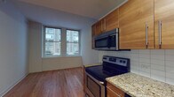 This single-floor urban residence is thoughtfully designed with modern finishes and a functional layout. The kitchen serves as a standout feature, equipped with warm wood cabinetry fitted with sleek bar-pull hardware, granite countertops, a white subway tile backsplash, and a full suite of stainless steel appliances including an electric range, over-the-range microwave, refrigerator, and dishwasher. A deep sink with a modern faucet completes the kitchen, which flows naturally toward the entry corridor finished with light hardwood flooring and recessed lighting. The living and sleeping areas feature a mix of hardwood flooring and carpeting, with clean white walls that create a bright and neutral backdrop. Large windows throughout the residence frame views of the surrounding urban streetscape and neighboring architectural facades, allowing natural light to filter into each room. The bedroom areas are generously proportioned with high ceilings, providing a spacious and airy feel. The overall design aesthetic is contemporary and understated, combining practical amenities with clean lines to create a comfortable urban living environment well-suited for city life.