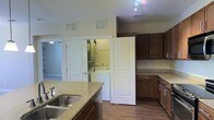 Located at 320 The Green Cir in Raleigh, NC, this single-floor residence offers a practical and well-appointed layout. The kitchen is a standout feature, showcasing rich dark wood cabinetry that extends to the upper and lower walls, providing ample storage. Granite-look countertops add a refined touch, complementing the stainless steel double-basin sink fitted with a modern faucet. A kitchen island mirrors the same countertop finish, offering additional prep space and functionality. The flooring throughout is a warm hardwood, lending a cohesive and inviting aesthetic to the space. Overhead track lighting in the kitchen ensures bright, focused illumination. A dedicated in-unit laundry area is conveniently accessible through a set of panel doors, housing a full-size washer and dryer side by side. An adjacent doorway leads to additional living space, where neutral-toned walls and consistent flooring continue the home's clean, understated design. Ceiling vents are thoughtfully placed throughout, supporting efficient climate control. Overall, the space presents a straightforward, functional living environment with quality finishes.