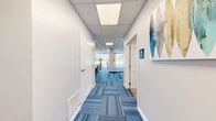 Located at 300 N Main St in Mansfield, Massachusetts, this single-floor fitness facility is thoughtfully designed to maximize functionality within a compact footprint. The space comprises a primary gym area, a hallway, a bathroom, and a closet, offering a complete and self-contained workout environment.

The gym is the centerpiece of the facility, featuring a distinctive blue multi-toned carpet tile flooring that adds a modern, energetic aesthetic throughout. Large floor-to-ceiling windows along one wall flood the space with abundant natural light and provide open views of the surrounding exterior, creating an airy and motivating atmosphere. The drop ceiling with recessed panel lighting ensures consistent, even illumination throughout.

The gym is equipped with a variety of fitness machines, including treadmills, an elliptical trainer, a multi-station cable machine, and a dumbbell rack with a full weight selection and a workout bench. A wall-mounted television and full-length mirrors further enhance the workout experience. Vibrant athletic artwork adorns the walls, contributing to the facility's energetic character.

The hallway provides clean, organized access between the gym, the closet for storage, and the accessible bathroom. The bathroom is finished in neutral tones with wood-look flooring, grab bars, and a paper towel dispenser, meeting accessibility standards. The overall design is clean, bright, and purpose-built for an effective fitness experience.