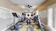 This fitness center offers a thoughtfully designed single-floor workout space with a practical and energizing layout. The room is anchored by a collection of modern cardio equipment, including treadmills, ellipticals, stationary bikes, and a rowing machine — all from Freemotion — arranged to maximize use of the available floor space. A distinctive feature of the room is its motivational word-art wall murals, which display fitness-themed typography in colorful text across multiple walls, creating an upbeat and encouraging atmosphere. The flooring consists of a patterned carpet tile in a geometric mix of yellow, gray, and tan tones, adding visual interest underfoot. Overhead, multiple ceiling fans provide consistent airflow throughout the space, complemented by fluorescent and recessed lighting for clear illumination. Natural light filters in through several windows fitted with blinds, as well as a set of glass French doors that open to the exterior. Additional amenities include a wall-mounted flat-screen television, a standing body weight scale, a hand sanitizer dispenser, and water coolers with bottle storage. A small adjacent area, visible through an interior doorway, appears to serve as an office or administrative nook. The space is practical, clean, and well-maintained, offering everything needed for a complete cardio-focused workout.