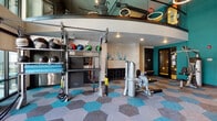 Located at 5745 Golden Triangle Blvd in Fort Worth, Texas, this two-story fitness center is designed to accommodate a wide range of workout styles and training needs. The space is immediately distinguished by its bold teal accent walls, eye-catching hexagonal patterned carpet in teal, blue, tan, and gray tones, and contemporary circular halo lighting fixtures suspended from above.

The main level offers an open, well-lit workout area featuring strength machines, rowing equipment, and Cybex cardio machines. A dedicated functional training zone houses a multi-use rack stocked with medicine balls, resistance bands, TRX suspension trainers, and a variety of accessories. Full-length mirrors line portions of the walls, enhancing the sense of space throughout. Large windows along one wall allow ample natural light to fill the floor, contributing to an energizing atmosphere.

A staircase connects the main level to the upper floor, which features additional open workout space, wall-mounted flat-screen televisions for guided training sessions, stability balls, and mirrored walls. The upper level overlooks the main floor through a partial open mezzanine design, reinforcing the facility's spacious, multi-dimensional layout. Exposed ductwork on the upper level adds an industrial design element that complements the overall modern aesthetic.