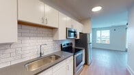 This single-floor residence presents a clean, modern design characterized by light hardwood-style flooring, crisp white walls, and contemporary finishes throughout. The galley-style kitchen is well-appointed, featuring white flat-front cabinetry with brushed metal hardware, dark countertops, and a white subway tile backsplash. A full suite of stainless steel appliances includes an electric range, over-the-range microwave, refrigerator, and dishwasher, offering a functional and cohesive cooking environment. Recessed and flush-mount ceiling lighting provides consistent illumination throughout the kitchen.

The open living area benefits from large windows fitted with horizontal blinds, allowing generous natural light to fill the space. A ceiling fan serves the main living area, adding both practical airflow and a subtle design element. The layout flows efficiently, connecting the kitchen and living spaces in a straightforward, open arrangement.

The bathroom is finished with a single-sink vanity topped with a dark countertop, white cabinetry, and a large framed mirror with modern bar lighting above. A tiled shower-and-tub combination completes the space. Overall, the unit offers a cohesive, contemporary aesthetic with practical, well-maintained finishes.