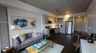 This single-floor space showcases a thoughtfully designed open-concept layout that seamlessly integrates the living and kitchen areas into one cohesive environment. The exposed concrete ceiling lends an industrial character to the space, while dark hardwood flooring grounds the interior with warmth and sophistication.

The kitchen is outfitted with sleek gray cabinetry, a classic white tile backsplash, and a full suite of stainless steel appliances, including a refrigerator, range, and microwave. The L-shaped counter configuration maximizes workspace and storage, making it both functional and visually streamlined. Track lighting overhead provides focused illumination across the cooking and prep areas.

The adjoining living area is bright and welcoming, anchored by a comfortable gray sofa paired with accent pillows and a stylish wood-and-metal coffee table. A geometric patterned artwork on the wall adds a bold visual focal point, complementing the contemporary aesthetic throughout. A textured area rug defines the seating zone, adding layers of comfort underfoot.

Together, the spaces create a clean, modern ambiance that balances industrial design elements with warm, residential touches — resulting in a unit that feels both functional and inviting.