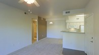 This single-floor residence offers a practical and functional open-concept layout that seamlessly connects the living area to the kitchen. The living space is finished with neutral-toned carpet and smooth ceilings, outfitted with a ceiling fan for comfort and a pendant light that adds a modest focal point overhead. A warm accent wall in taupe provides a subtle visual contrast against the lighter surrounding walls, adding dimension to the space.

The kitchen is positioned along one side of the open floor plan and features white cabinetry, a built-in microwave, and a raised breakfast bar countertop that doubles as a casual dining or serving surface. The counter material offers a contrasting tone to the white cabinetry, creating a clean and cohesive look. Overhead vents are strategically placed throughout, ensuring consistent climate control across the space.

A hallway visible from the main living area leads to additional rooms, hinting at a thoughtfully laid-out floor plan. The glimpse into the adjoining space reveals continued light-toned finishes, maintaining a consistent aesthetic throughout. The overall ambiance of the residence is bright, clean, and move-in ready, making it well-suited for those seeking a straightforward and comfortable living environment.