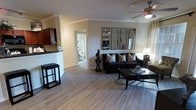 This single-story residence showcases an open-concept layout that connects the living room and kitchen into one cohesive, functional space. The living area features light-toned hardwood-style flooring that runs throughout, complementing the neutral beige walls adorned with crown molding for a polished finish. A ceiling fan with an integrated light fixture serves as a practical central focal point overhead.

The living room is anchored by a dark-toned sofa paired with a matching coffee table, while a set of four decorative geometric mirror panels adds visual depth and dimension to the wall. A floor lamp provides warm accent lighting alongside the natural light that fills the room.

Adjacent to the living area, the kitchen is partially visible and features rich dark cabinetry with a raised breakfast bar, ideal for casual dining or entertaining. A bar stool positioned at the counter adds a functional design element.

A doorway leading to a bathroom is visible from the main living space, offering a glimpse of the home's thoughtful layout. Overall, the space carries a contemporary, well-appointed ambiance with cohesive dark and neutral tones throughout.