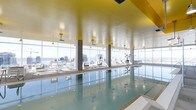 This indoor pool facility is a striking amenity space defined by its dramatic floor-to-ceiling glazed walls, which wrap around the perimeter and flood the interior with abundant natural light. The expansive windows frame panoramic views of the surrounding cityscape and what appears to be a waterfront, creating a visually immersive environment for swimmers and loungers alike.

The pool itself is generously sized, with calm, clear water that reflects the bright surroundings. A smooth pool deck lines the length of the pool, furnished with sleek lounge chairs arranged to take full advantage of the views. Structural columns punctuate the space at regular intervals, subtly framing the outlook without obstructing it.

One of the most distinctive architectural features is the bold yellow ceiling, which adds a warm, energetic contrast to the cool tones of the water and the neutral finishes of the walls and flooring. Pendant and wall-mounted light fixtures supplement the natural light, ensuring the space remains well-lit at all times.

Overall, the space conveys a refined, resort-like ambiance, combining functional aquatic amenities with thoughtful design elements that prioritize light, views, and visual comfort.