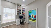 This well-designed single-floor space seamlessly blends productivity and play in a bright, welcoming environment. The main office area features a built-in white shelving unit with a granite-topped desk, offering ample storage for books and decorative items. Light hardwood-style flooring, neutral gray walls, and large windows fitted with horizontal blinds allow natural light to fill the room. A set of elegant French doors with glass panels adds architectural character and connects the space to an adjacent area.

Directly accessible from the office is a vibrant children's playroom that bursts with color and imagination. The room is painted in bold turquoise with large, hand-painted flower murals adorning the walls and decorative oversized leaf panels hanging from the ceiling, creating an immersive garden-inspired environment. Soft animal-shaped seating and a child-sized play table complete the space.

A hallway leads to a practical refreshment nook featuring white cabinetry, a marble-style countertop with subway tile backsplash, and dual water fountains mounted conveniently in the corridor. A restroom is also accessible nearby, rounding out the functional amenities. Recessed lighting throughout ensures a consistently well-lit and inviting atmosphere across all areas.