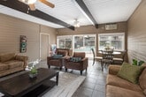 Located in Orange, California, this single-story clubhouse offers a warm and inviting gathering space defined by its distinctive architectural character. The room is lined with horizontal wood-paneled walls in a soft tan tone, lending a casual, retro charm throughout. Exposed dark beams span the white-paneled ceiling, adding visual depth and a rustic touch, while a ceiling fan ensures year-round comfort. A classic full-brick fireplace serves as the room's focal centerpiece, framed by windows on either side that bring in generous natural light and views of the surrounding neighborhood. The durable tile flooring grounds the space with a practical yet attractive finish. On the opposite end of the room, a wall of sliding glass doors and large windows opens the interior to direct views of the outdoor pool area, seamlessly connecting the indoor lounge to the community's exterior amenities. A dining area with a glass-top table and chairs sits adjacent to this pool-facing wall, providing a functional space for casual meals or social gatherings. A wall-mounted book rack and additional seating nooks offer quiet retreats within the room. Overall, the clubhouse presents a comfortable, community-oriented environment well-suited for socializing and relaxation.