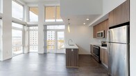 This contemporary single-floor residence showcases a thoughtfully designed open-concept layout with striking architectural details throughout. The living and kitchen area is anchored by soaring double-height ceilings and floor-to-ceiling windows that flood the space with natural light and frame expansive urban and treetop views. A private balcony is accessed through large glass doors, extending the living space outdoors.

The modern kitchen features warm wood-toned flat-front cabinetry, a subway tile backsplash, and a full suite of stainless steel appliances including a range, microwave, and refrigerator. A central island with a crisp white countertop and integrated sink serves as both a functional workspace and a casual gathering point. Pendant lighting adds a refined touch overhead. Dark hardwood-style flooring runs throughout the main living areas, providing a grounding contrast to the bright, airy palette.

The bedrooms are carpeted for comfort and benefit from large windows with urban outlooks. Each bedroom includes access to a closet or walk-in closet with wire shelving. The bathrooms are finished to a high standard, featuring floating vanities with white countertops, backlit mirrors, and dark wood-style flooring. One bathroom includes a soaking tub with a tiled surround.