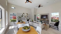 This single-story space is defined by its dramatic vaulted ceilings and open-plan layout, creating a spacious, airy atmosphere throughout. Light-toned tile flooring and crisp white walls reflect natural light that pours in from an abundance of large windows, offering views of lush tropical greenery and an outdoor patio area beyond.

The living area is generously proportioned, accommodating a comfortable sectional sofa, accent seating, and a wall-mounted television framed within a recessed niche. Multiple ceiling fans with wood-toned blades provide both function and a casual, coastal aesthetic. A set of glass-paneled double doors provides direct access to the outdoors, reinforcing the connection between interior and exterior spaces.

Adjacent to the living area, a dedicated dining space features a wood-topped table with modern white chairs, bathed in natural light from floor-to-ceiling windows overlooking the surrounding landscape. Clerestory windows above add to the sense of height and brightness.

A compact kitchenette occupies one wall, outfitted with white cabinetry, a tile backsplash, open shelving, a microwave, mini-refrigerator, and sink. The overall design palette is clean and contemporary, combining neutral tones with modern fixtures to create a welcoming, versatile environment suited for gathering and socializing.
