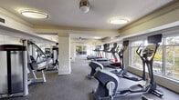 This fitness center is a comprehensively equipped facility designed to accommodate a wide range of workout routines. The space stretches across a single floor, offering an elongated layout that provides ample room for both cardio and strength training activities. Structural columns are positioned throughout, defining distinct workout zones without impeding movement or sight lines.

Along one side of the facility, a row of large windows allows natural light to fill the space, offering views of the surrounding exterior. Opposite the windows, floor-to-ceiling mirrors line the walls, visually expanding the room and allowing users to monitor their form during exercise. The mirrors also reflect the natural light, contributing to a bright and open atmosphere.

The flooring consists of durable, speckled grey carpet suited for fitness use, while the warm yellow-toned walls and white crown molding lend a clean, polished aesthetic. Flush-mount and pendant lighting fixtures are evenly distributed across the ceiling, ensuring consistent illumination throughout.

The cardio section features multiple treadmills, elliptical trainers, and stationary bikes from FreeMotion, arranged in orderly rows. A dedicated strength training area includes cable machines and resistance equipment, while a free weight station with a full dumbbell rack occupies a separate zone. A wall-mounted television provides entertainment, completing this well-rounded fitness environment.
