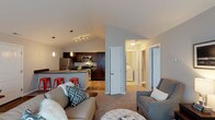 This single-floor residence showcases an open-concept layout that seamlessly connects the living and kitchen areas. The kitchen is equipped with stainless steel appliances, including a refrigerator, microwave, and range, complemented by rich dark cabinetry and a peninsula countertop with bar seating accommodated by red metal stools. Pendant lighting fixtures hang above the peninsula, adding both function and style, while a track lighting system illuminates the kitchen workspace. The vaulted ceilings throughout enhance the sense of spaciousness and airiness. The living area features plush carpeting and is furnished with comfortable seating, creating a welcoming atmosphere. A hallway leads to additional spaces, including an in-unit laundry room with a side-by-side washer and dryer, and a walk-in closet offering generous storage. Doors off the hallway suggest access to a bedroom and bathroom. The overall design balances practicality with modern aesthetics, featuring a neutral color palette of whites and grays accented by warm wood tones in the cabinetry and light wood flooring in the kitchen.