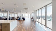 Located on the upper floors of 1 Commercial Place in Norfolk, VA, this generously sized amenity space offers an impressive open-plan layout spanning a single floor. Rich, multi-toned hardwood flooring runs throughout, lending warmth and continuity to the expansive interior. Floor-to-ceiling glass walls and sliding doors line multiple sides of the space, flooding the interior with abundant natural light and framing sweeping panoramic views of the Norfolk skyline and surrounding waterfront. The open layout is thoughtfully organized into distinct functional zones, including multiple seating lounges furnished with plush blue and gray velvet sofas and accent chairs, as well as a casual dining and bar area with counter seating. Gold-toned open shelving units serve as elegant dividers between zones, adding a refined, contemporary touch. A wall-mounted flat-screen television anchors one of the lounge areas, making it well-suited for gatherings and presentations. Recessed lighting and pendant fixtures complement the natural light throughout. Slender white columns punctuate the space without interrupting sightlines. Access to an outdoor terrace further enhances the experience, allowing residents or guests to take in unobstructed elevated views of the city and beyond. The overall ambiance is modern, sophisticated, and airy.