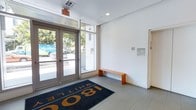 This single-floor lobby space at 1801 Whittle offers a crisp, modern first impression with a distinctly minimalist design. The entry is anchored by a set of full-length glass double doors framed in brushed metal, allowing abundant natural light to flood the interior and providing clear sightlines to the lush greenery outside. Transom windows above the doors further enhance the brightness of the space.

The flooring features smooth, light-toned tiles that extend throughout the lobby, contributing to a clean and polished look. A bold branded entrance mat bearing the "1801 Whittle" name is prominently placed near the entry, reinforcing the building's identity. The walls are finished in a bright white, lending an open and airy quality to the space.

A simple orange bench is positioned along one wall, offering a functional seating option for visitors and residents. Subtle architectural details, including recessed ceiling lighting and a neatly mounted evacuation plan and intercom panel on the wall, reflect thoughtful and practical design. A doorway on the adjacent wall provides access to the building's interior. Overall, the lobby communicates a professional, well-maintained environment with a contemporary character.