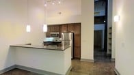 This single-floor space showcases an open, loft-inspired design characterized by dramatically high ceilings that create an airy, expansive atmosphere. The polished concrete flooring runs throughout, lending an industrial aesthetic that pairs well with the clean white walls.

The kitchen is a central feature, equipped with stainless steel appliances including a refrigerator, electric range, and over-the-range microwave. Warm-toned wood cabinetry provides generous storage, extending from counter height to near the ceiling. A peninsula with a dark granite countertop offers additional workspace and defines the kitchen area within the open layout.

Overhead, a track lighting system illuminates the kitchen, complemented by pendant lights that add a modern touch. An adjacent open area, visible beyond the kitchen, continues the concrete flooring and high ceilings, suggesting flexible use of space. The overall ambiance balances industrial elements—exposed structural details, raw concrete—with refined finishes, making the space versatile for a variety of functional arrangements.