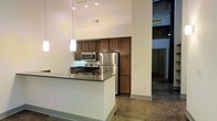 This single-floor space showcases an open, loft-inspired design characterized by dramatically high ceilings that create an airy, expansive atmosphere. The polished concrete flooring runs throughout, lending an industrial aesthetic that pairs well with the clean white walls.

The kitchen is a central feature, equipped with stainless steel appliances including a refrigerator, electric range, and over-the-range microwave. Warm-toned wood cabinetry provides generous storage, extending from counter height to near the ceiling. A peninsula with a dark granite countertop offers additional workspace and defines the kitchen area within the open layout.

Overhead, a track lighting system illuminates the kitchen, complemented by pendant lights that add a modern touch. An adjacent open area, visible beyond the kitchen, continues the concrete flooring and high ceilings, suggesting flexible use of space. The overall ambiance balances industrial elements—exposed structural details, raw concrete—with refined finishes, making the space versatile for a variety of functional arrangements.