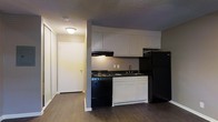 This single-floor space features a practical, open-concept kitchen that serves as a central element of the layout. The kitchen is equipped with black appliances, including a range, overhead range hood, and a full-sized refrigerator. White upper and lower cabinetry provides ample storage, paired with dark countertops that offer a contrasting visual accent. A single-basin sink is positioned beneath the upper cabinets, keeping the workspace efficient and organized.

The flooring throughout is a dark wood-style laminate or vinyl, lending warmth and continuity to the space. A textured ceiling is present overhead, with a flush-mount ceiling light providing ambient illumination. The walls are finished in a neutral tone, contributing to a clean and understated aesthetic.

Adjacent to the kitchen, a hallway leads to what appears to be a closet with a sliding door and a separate entry door, suggesting additional functional zones within the unit. The overall layout is compact yet efficient, designed to maximize utility within the available footprint. The space presents a straightforward, move-in-ready aesthetic suitable for practical everyday living.