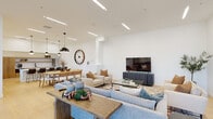 This thoughtfully designed single-floor space combines modern aesthetics with functional communal living. The entrance greets visitors with a bright, airy lobby featuring floor-to-ceiling black-framed glass doors that flood the space with natural light and offer views of the surrounding greenery. A sculptural dark wood accent table anchors the entryway, setting an elegant tone throughout.

The open-plan interior flows seamlessly from the lobby into a generous lounge and dining area. The lounge is anchored by a large sectional sofa, accent chairs, and a glass-top coffee table arranged over a richly patterned area rug, all centered around a wall-mounted flat-screen television on a navy media console. A striking oversized wooden clock serves as a bold focal point on the feature wall, adding a distinctive design element to the dining zone, which accommodates a long communal wooden table beneath a dramatic pendant light.

The kitchen is sleek and contemporary, outfitted with warm wood cabinetry, white quartz countertops, subway tile backsplash, open shelving, and stainless steel appliances. Globe and dome pendant lights hang above a generously sized island with bar-height seating. A separate casual seating nook, adorned with minimalist abstract artwork, provides additional space for informal gatherings. Light hardwood flooring and recessed linear ceiling lighting run throughout, contributing to the space's cohesive, modern ambiance.