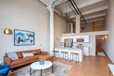 Located at 280–298 N Third St in downtown St. Joseph, Missouri, this single-floor loft showcases a compelling blend of historic industrial architecture and modern interior finishes. The open-concept living area commands immediate attention with its dramatically tall ceilings, original structural columns, and exposed ductwork running overhead, all accented by a beamed ceiling that nods to the building's historic character. Light hardwood flooring flows continuously throughout the space, reinforcing a sense of cohesion and warmth.

The living area is anchored by a large arched window with a bold dark frame, allowing abundant natural light to fill the room while offering views of the surrounding downtown streetscape. The kitchen features white shaker cabinetry extending toward the ceiling, granite-style countertops, and a full suite of stainless steel appliances including a refrigerator, range, microwave, and dishwasher. A peninsula with bar seating connects the kitchen seamlessly to the living space.

The bathroom is finished with white cabinetry, a solid white countertop vanity, and matte black fixtures, providing a clean, contemporary aesthetic. A dedicated utility area houses an in-unit washer and dryer alongside the water heater and HVAC system. The overall ambiance is urban and sophisticated, balancing raw architectural character with refined, modern detailing throughout.