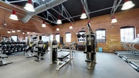 This gym occupies a striking industrial-style space that blends historic architectural character with modern fitness functionality. The exposed brick walls, darkened ceiling, and visible overhead ductwork and pipes give the space a rugged, warehouse-inspired aesthetic. Rows of pendant lights with bell-shaped shades hang from above, casting bright, even illumination throughout the training floor. The soaring ceiling height creates an open, airy atmosphere that adds to the sense of spaciousness.

The workout floor is finished with durable rubber flooring, providing both safety and practicality underfoot. A comprehensive array of strength training machines is arranged throughout the space, including cable systems, resistance machines, and benches suited for a variety of exercises. Along the perimeter, additional equipment such as free weights and supplementary training tools are neatly organized. A row of arched windows with dark frames lines the brick walls, allowing natural light to filter in and enhancing the overall ambiance. The combination of industrial design elements and well-maintained equipment makes this gym a functional and visually distinctive training environment.
