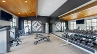 Located at 10999 SE 37th Ave in Milwaukie, Oregon, this well-appointed fitness center offers a generous, single-floor layout designed to support a full range of workout styles. The space is defined by its striking architectural detail — a warm, natural wood plank ceiling that spans much of the facility, complemented by exposed beam accents and recessed lighting that create an inviting yet energizing atmosphere.

The main workout floor is divided into distinct functional zones. The cardio area lines the perimeter, featuring treadmills, ellipticals, stair climbers, and rowing machines positioned alongside large multi-pane windows that flood the space with natural light and offer views of the surrounding neighborhood. Wall-mounted televisions are stationed throughout for entertainment during workouts.

The strength training zone is anchored by a bold design aesthetic — deep charcoal-colored accent walls adorned with geometric tile artwork serve as a dramatic backdrop for cable machines, a functional trainer, adjustable benches, and a full rack of dumbbells. Floor-to-ceiling mirrors amplify the sense of space and support proper form.

A glass-walled studio area provides a dedicated space for on-demand fitness programming, visible from the main floor entry. The entry corridor itself is clean and modern, featuring glass partitions and direct access to the exterior. Durable, neutral-toned carpet tile flooring ties the entire space together, making it both practical and polished.