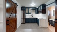 This single-floor space combines modern design elements with rustic touches to create a functional and visually distinctive layout. The open floor plan is anchored by light gray tile flooring that runs throughout, providing a clean, cohesive foundation across all areas.

The kitchen is a standout feature, showcasing deep teal shaker-style cabinetry with glass-front upper cabinets and chrome hardware. A classic white subway tile backsplash contrasts beautifully against the bold cabinetry, while a marble-look quartz countertop adds an elegant finish. Stainless steel appliances, including a French door refrigerator and microwave, complete the well-appointed cooking space. A rustic barn door on a black sliding hardware track serves as a charming and space-efficient entryway into the kitchen.

The living area is bright and open, with neutral gray walls, crown molding, and a modern flush-mount ceiling fan with integrated lighting. A wood-framed window allows natural light to filter in, while a wall-mounted HVAC unit provides climate control.

The bathroom features a frameless glass-enclosed walk-in shower dressed in rich dark stone-look tile, a rainfall showerhead, and a handheld fixture. A contemporary glass vessel sink rests atop a marble-surface vanity, adding a refined, modern touch. The overall ambiance of the space is clean, contemporary, and thoughtfully designed.