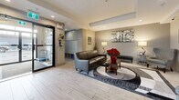Located at 691 Hoylake Ave in Langford, British Columbia, this thoughtfully designed lobby serves as an inviting gateway for residents and visitors alike. The space is finished with light wood-look tile flooring throughout, lending a clean and contemporary aesthetic that carries seamlessly across the entire area.

The main seating lounge is anchored by a tufted leather sofa and coordinating armchairs arranged atop a bold geometric area rug, creating a comfortable and welcoming gathering point. Warm table lamps, framed artwork, and fresh floral accents add a refined, homey character to the space.

A dedicated mailbox alcove is neatly integrated near the building entrance, providing convenient parcel and mail access for residents. Adjacent to this area, a glass-paneled elevator offers accessible vertical circulation within the building.

Further into the lobby, a secondary lounge area features a wingback accent chair, a decorative fireplace, lush indoor plants, and a stylish credenza topped with a sunburst mirror and a welcoming display — all contributing to a warm and curated ambiance. Bold, large-format artwork punctuates the walls throughout, adding visual energy and personality.

Natural light filters in through generous windows, brightening the space and offering views to the exterior streetscape. The overall design balances practicality with polished, modern interior styling, making for a memorable and functional building entrance.