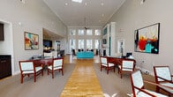 Located at 2067 Springdale Ln in Gallatin, Tennessee, this single-floor community or leasing facility offers an expansive, well-designed interior that blends professional functionality with inviting comfort. The main gathering area is defined by dramatically soaring ceilings and an abundance of natural light streaming through large, multi-pane windows and clerestory glazing above. Warm hardwood flooring runs throughout the open central corridor, anchored by a vibrant teal sofa and seating arrangements that create a welcoming lounge atmosphere. A ceiling fan and recessed lighting ensure year-round comfort and consistent illumination. Adjacent to the lounge, a fully equipped kitchen features dark cabinetry, a center island with bar seating, pendant lighting, and French door access to an outdoor area. The space also includes multiple private office areas, each furnished with glass-top executive desks, upholstered guest chairs, and large windows fitted with blinds for adjustable privacy. Colorful abstract artwork adds energy and personality to the office walls. A dedicated conference nook with a round glass table provides an intimate setting for small meetings, complemented by a nearby built-in workstation housing office equipment. Crown molding and clean neutral tones throughout lend the space a polished, professional character.