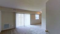 This single-floor residential space offers a clean and functional layout designed for comfortable everyday living. The main living area is a standout feature, characterized by its generous open floor plan and an abundance of natural light streaming in through large sliding glass doors dressed with full-length vertical blinds. A secondary window further brightens the space, creating a warm and airy atmosphere throughout. The floors are covered in soft, plush wall-to-wall carpeting in a neutral tone, contributing to the overall sense of warmth and comfort. The walls are finished in a light beige palette, lending the space a calm and inviting ambiance. Ceiling-mounted lighting provides consistent illumination, complementing the natural light sources. A visible HVAC vent in the ceiling ensures climate control throughout the living area. The space maintains a simple, uncluttered aesthetic with smooth walls and standard interior doors, making it well-suited for a variety of furniture arrangements and personal styles. Overall, this single-floor space presents a practical and livable environment with a neutral, move-in-ready interior.
