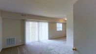 This single-floor residential space offers a clean and functional layout designed for comfortable everyday living. The main living area is a standout feature, characterized by its generous open floor plan and an abundance of natural light streaming in through large sliding glass doors dressed with full-length vertical blinds. A secondary window further brightens the space, creating a warm and airy atmosphere throughout. The floors are covered in soft, plush wall-to-wall carpeting in a neutral tone, contributing to the overall sense of warmth and comfort. The walls are finished in a light beige palette, lending the space a calm and inviting ambiance. Ceiling-mounted lighting provides consistent illumination, complementing the natural light sources. A visible HVAC vent in the ceiling ensures climate control throughout the living area. The space maintains a simple, uncluttered aesthetic with smooth walls and standard interior doors, making it well-suited for a variety of furniture arrangements and personal styles. Overall, this single-floor space presents a practical and livable environment with a neutral, move-in-ready interior.