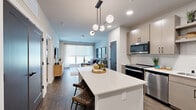 This 1-bedroom, 1-bathroom residence in Milwaukee, WI offers a modern, open-concept layout that makes efficient use of its well-proportioned footprint. The combined kitchen and living area serves as the heart of the home, anchored by a sleek white quartz island with built-in shelving and barstool seating, illuminated by a contemporary globe pendant light fixture. The kitchen features light wood cabinetry, a white subway tile backsplash, stainless steel appliances including a refrigerator, range, microwave, and dishwasher, and a deep single-basin sink. Rich wood-tone flooring flows throughout the main living area, complementing the neutral palette. The living room is bright and inviting, bathed in natural light from a large picture window with sheer drapery. The bedroom offers plush carpeting, a generous walk-in closet, and a large window with outdoor views. Three closets total provide ample storage throughout the unit. Additional features include a full bathroom, an in-unit laundry area, and a private patio. The overall design balances contemporary aesthetics with functional living in a clean, well-appointed space.